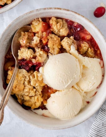 A white bowl of cranberry apple crisp with a spoon and topped with scoops of vanilla ice cream.
