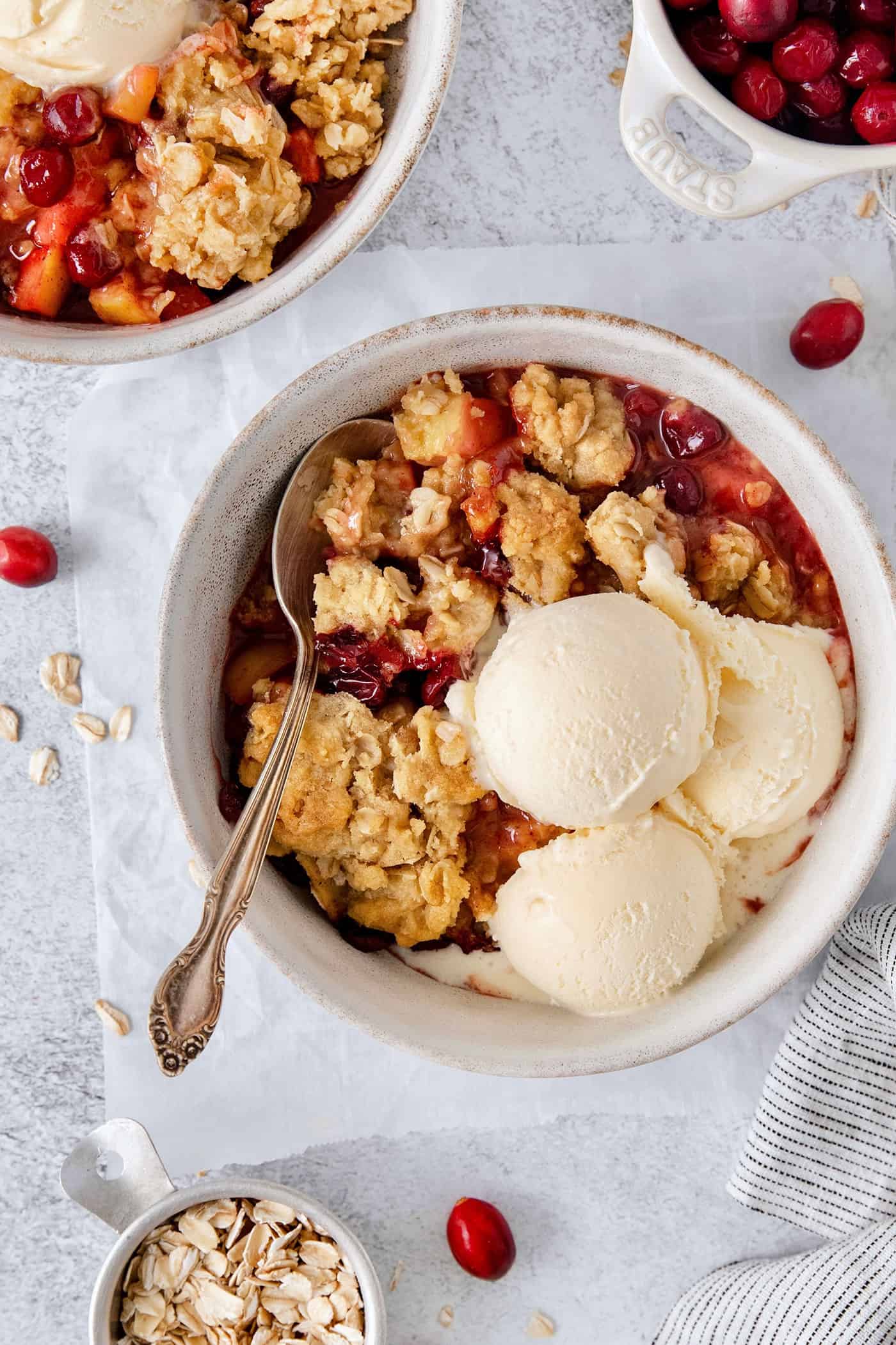 This Cranberry Apple Crisp features fresh, sweet-tart cranberries and apples, plus a crispy, buttery oat topping. A white bowl of cranberry apple crisp with a spoon and topped with scoops of vanilla ice cream.
