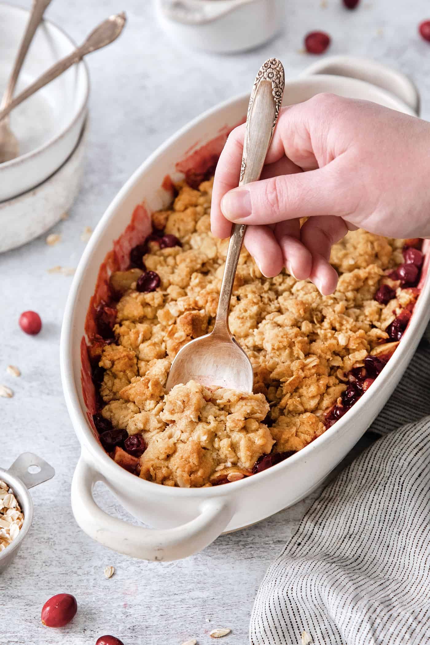 This Cranberry Apple Crisp features fresh, sweet-tart cranberries and apples, plus a crispy, buttery oat topping. A hand holding a spoon cuts into a dish of cranberry apple crisp.