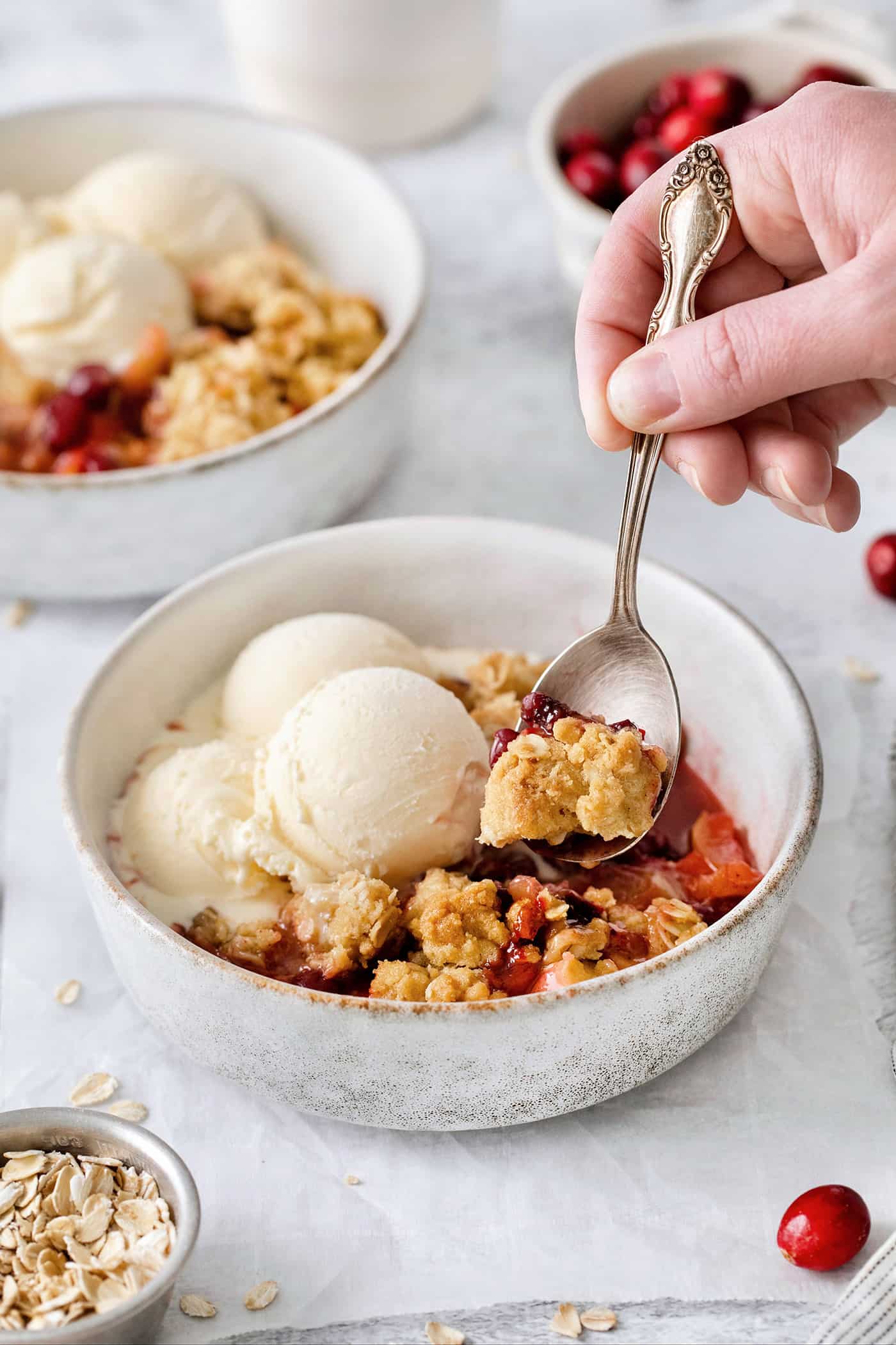 This Cranberry Apple Crisp features fresh, sweet-tart cranberries and apples, plus a crispy, buttery oat topping. A hand holding a spoon dips into a bowl of cranberry apple crisp topped with vanilla ice cream.