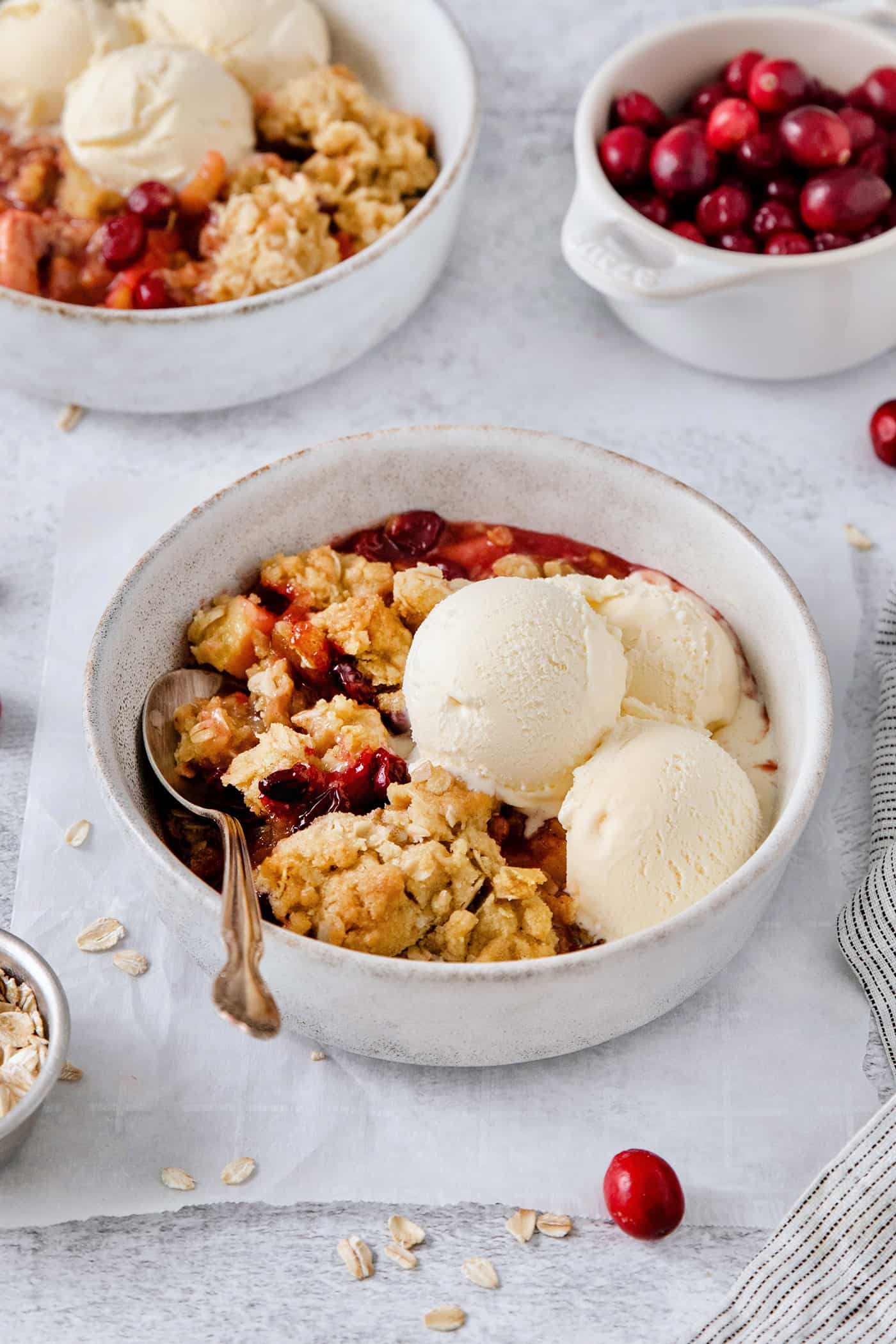 This Cranberry Apple Crisp features fresh, sweet-tart cranberries and apples, plus a crispy, buttery oat topping. A white bowl of cranberry apple crisp with a spoon and topped with scoops of vanilla ice cream.