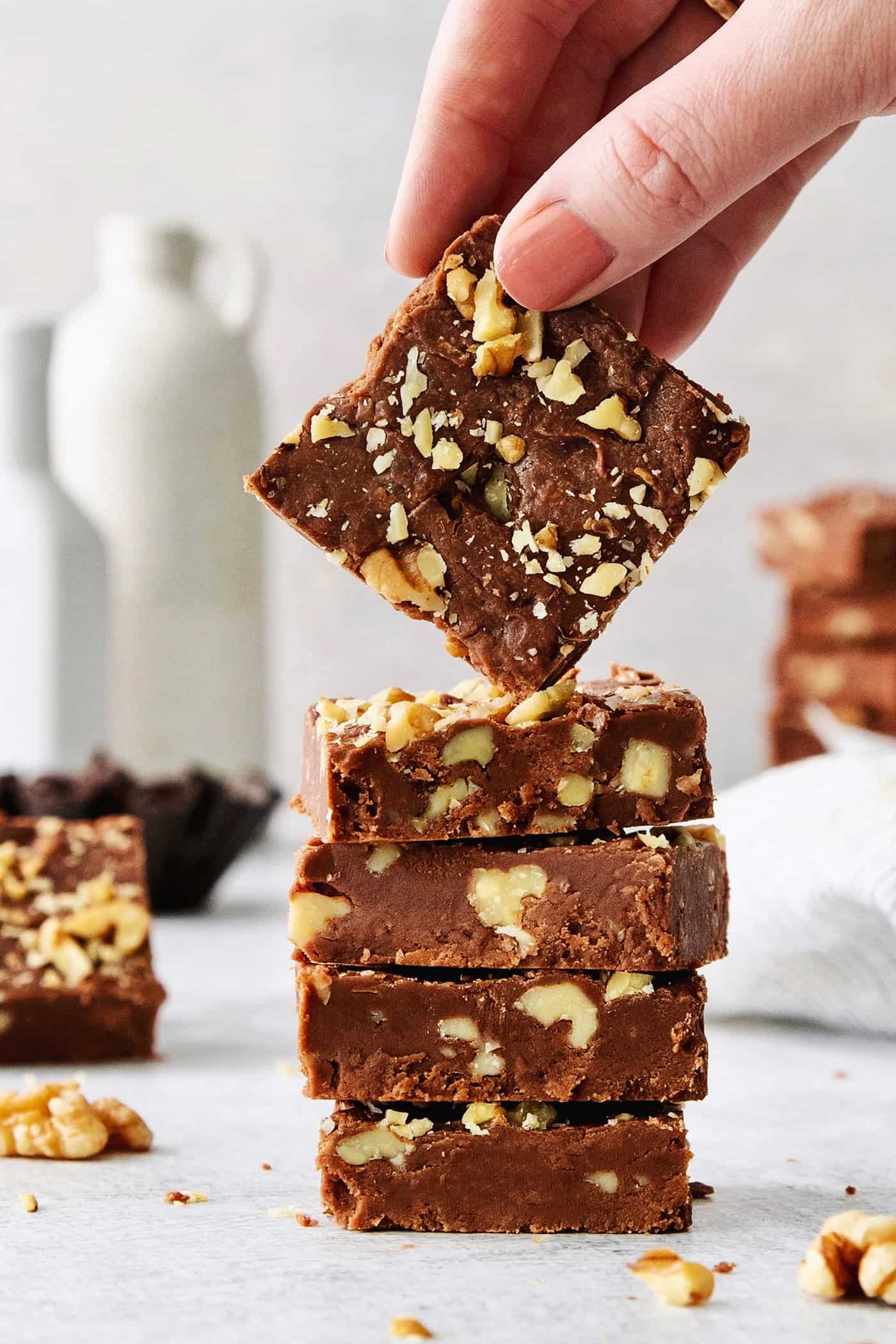 Fantasy Fudge is a thick, rich, no-fail chocolatey fudge made with semi-sweet chocolate, marshmallow creme, and walnuts. A hand lifts a piece of Fantasy Fudge off the top of a stack of fudge.