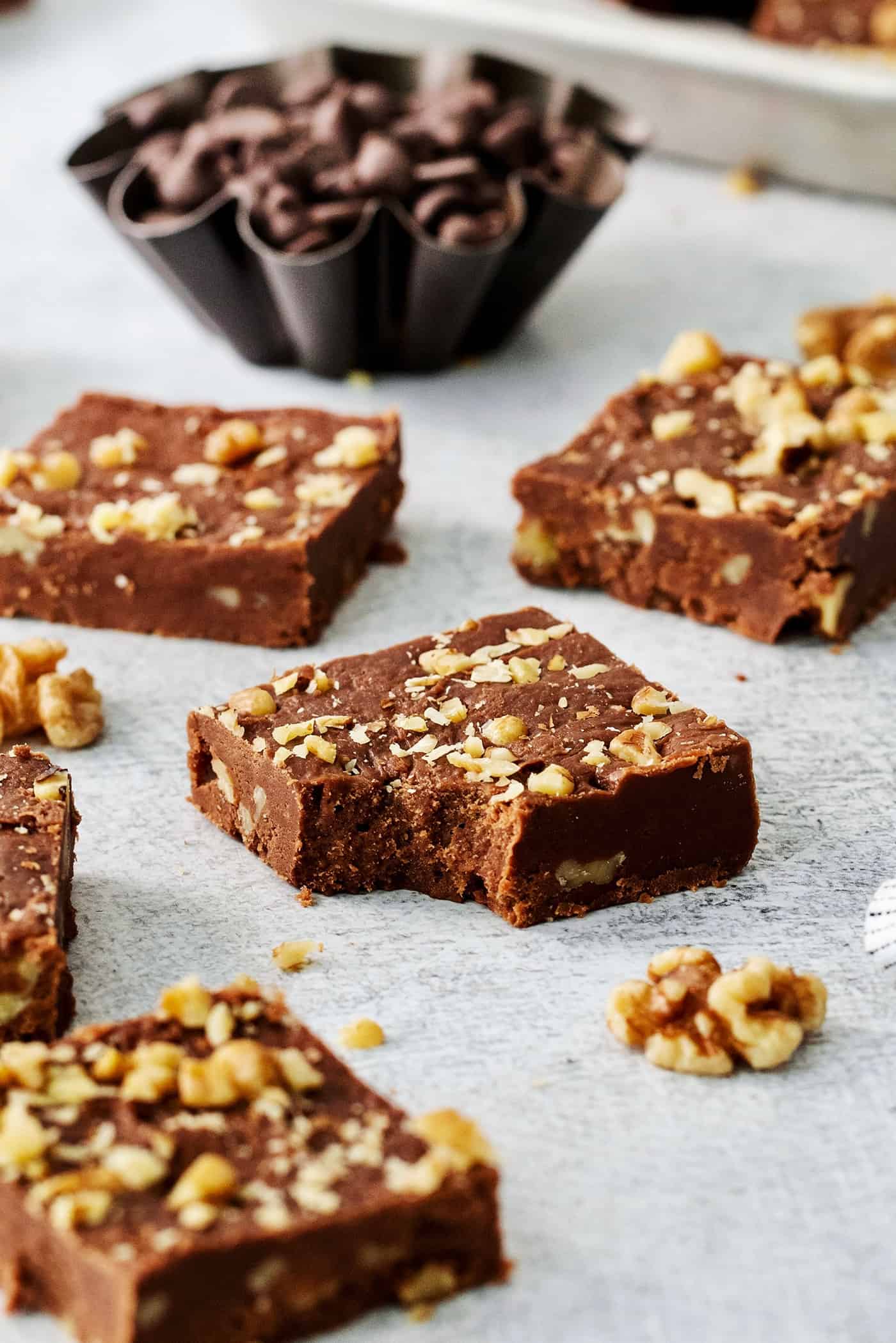 Fantasy Fudge is a thick, rich, no-fail chocolatey fudge made with semi-sweet chocolate, marshmallow creme, and walnuts. Pieces of Fantasy Fudge are shown on a white background with a bite taken out of a piece.
