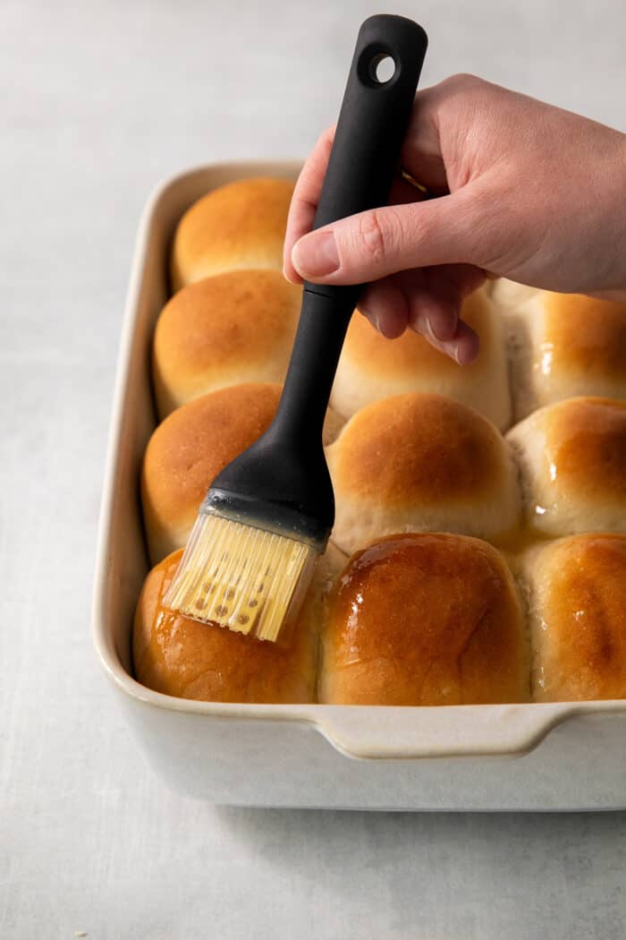 These copycat Rhodes Rolls are light and fluffy with a tender crumb and delicate flavor. Perfect for holiday meals! A hand holding a pastry brush brushes butter on a pan of rhodes rolls.