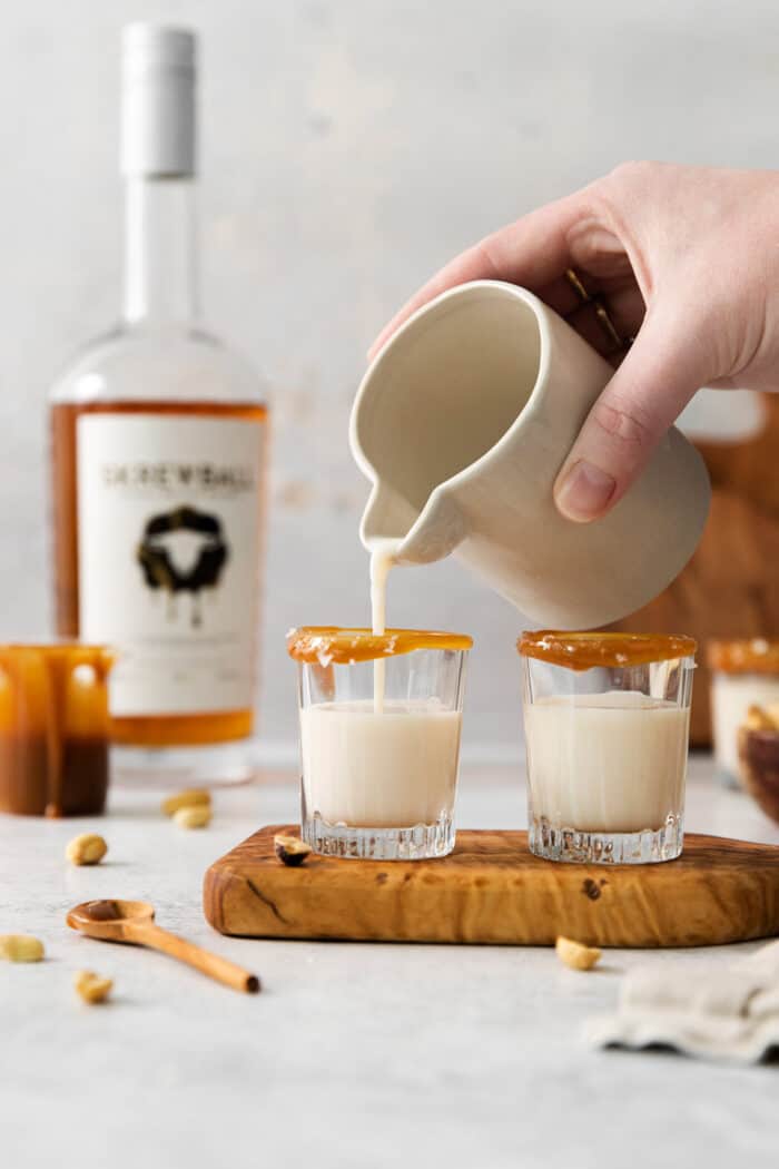 This Salted Nut Roll Shot is a fantastic twist on the salted nut roll candy bar with Rumchata, peanut butter whiskey, and vanilla vodka. A hand holding a pitcher pours salted nut roll shots into shot glasses.