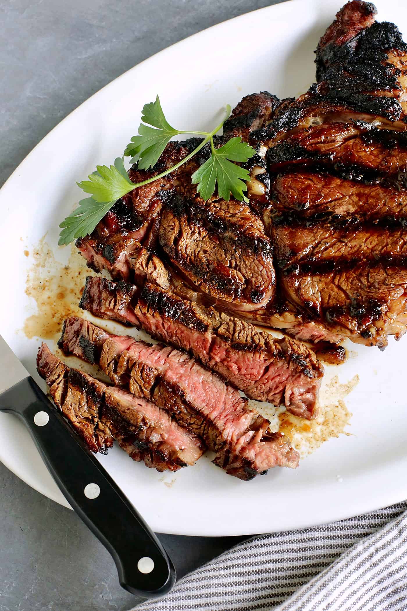 Elevate steak night and make Sugar Grilled Steak! Sprinkled generously with sugar, the grill's fire caramelizes the meat to juicy perfection. A platter of sliced sugar grilled steak with a knife resting on the edge of the plate.