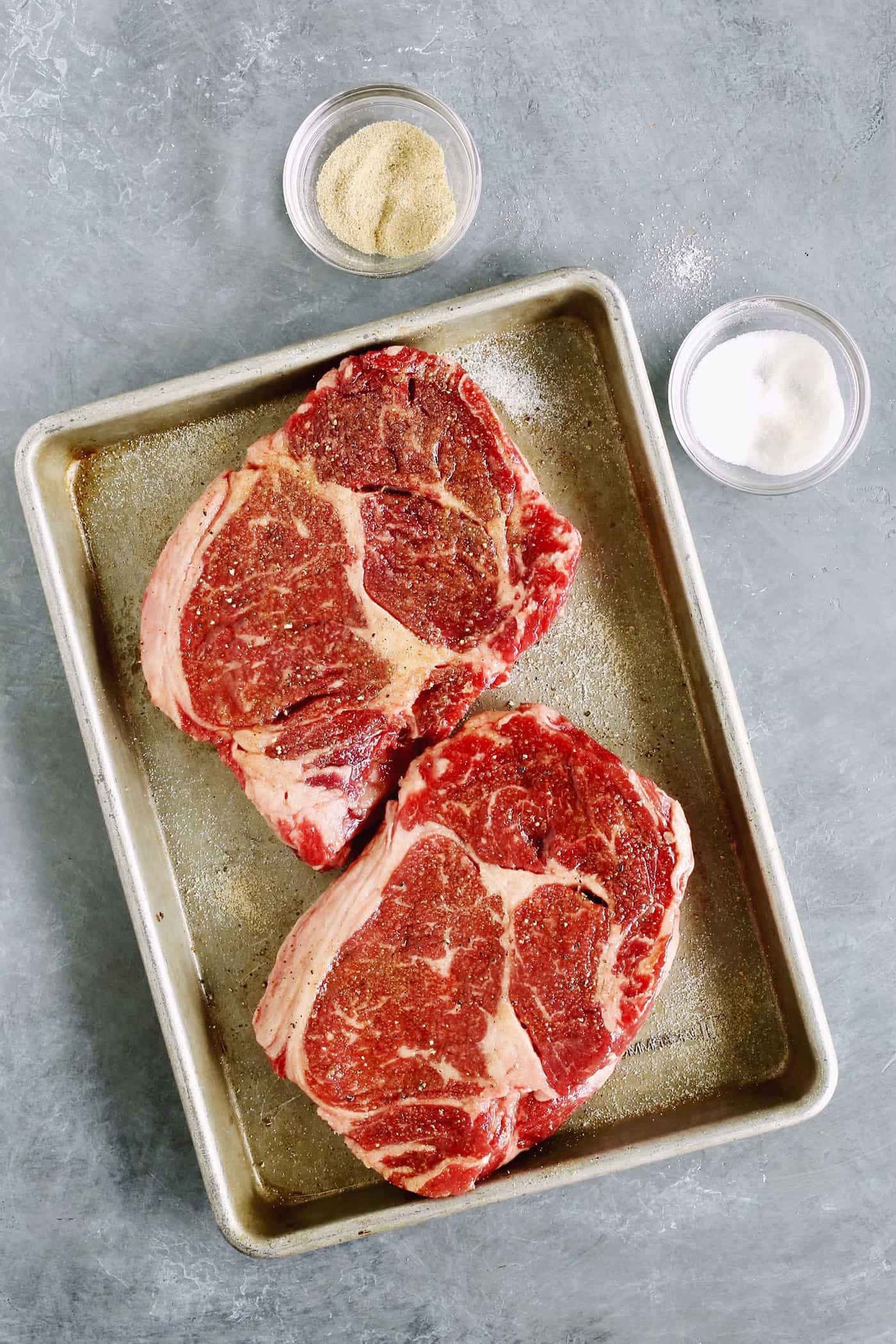 Elevate steak night and make Sugar Grilled Steak! Sprinkled generously with sugar, the grill's fire caramelizes the meat to juicy perfection. Two steaks are shown on a baking sheet with salt and sugar next to it.