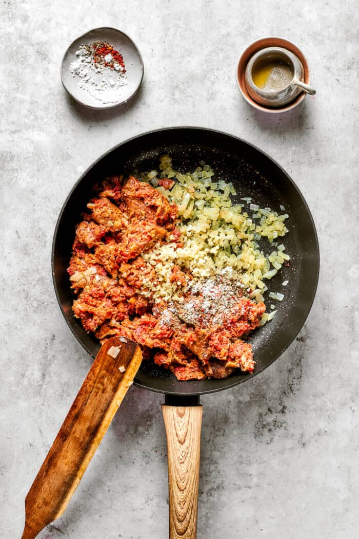 These cheesy Lasagna Roll Ups include a hearty blend of cheeses and sausage-flecked meat sauce that's wrapped in tender noodles. Garlic, onion, and sausage cook in a black skillet, being stirred by a wooden spoon, with small bowls of seasonings nearby.