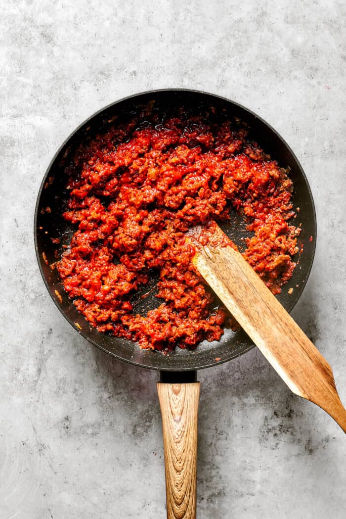 These cheesy Lasagna Roll Ups include a hearty blend of cheeses and sausage-flecked meat sauce that's wrapped in tender noodles. A wooden stirring paddle stirs red sauce and meat together in a black skillet.