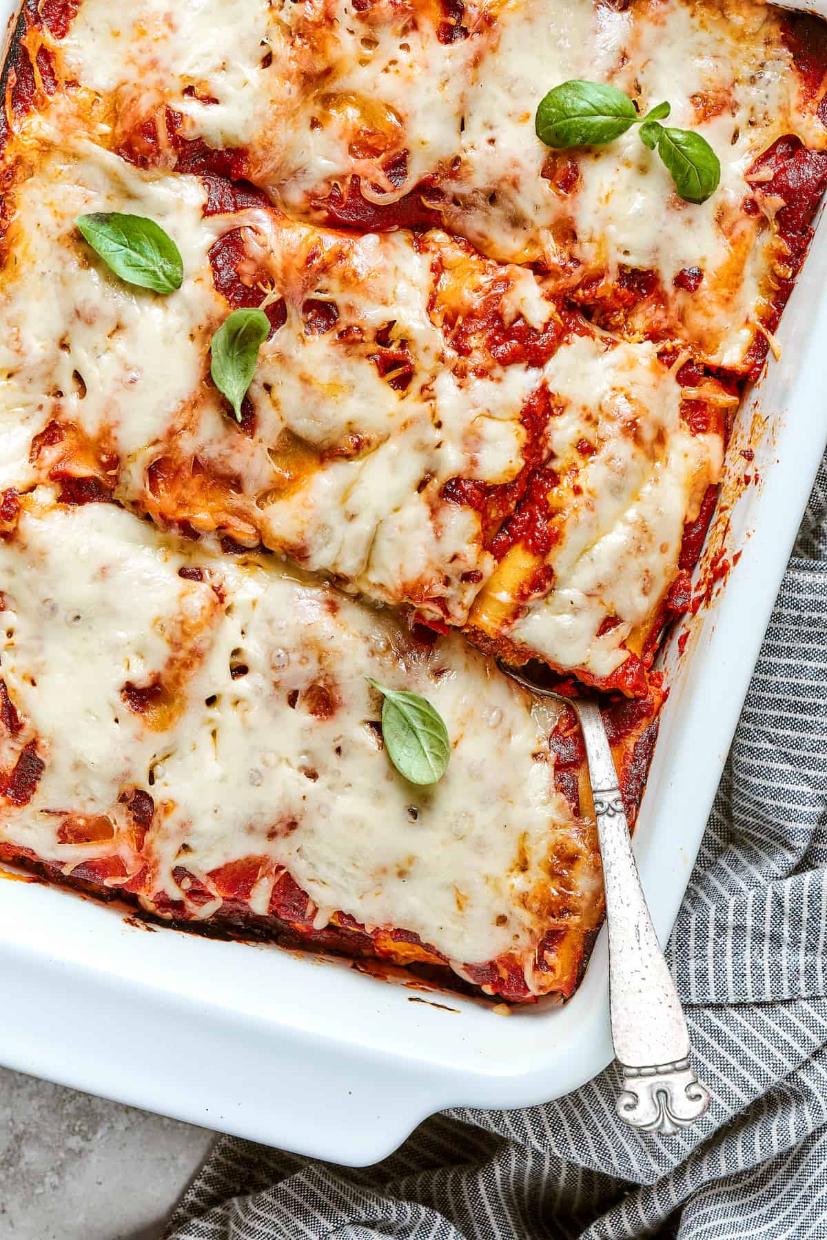 These cheesy Lasagna Roll Ups include a hearty blend of cheeses and sausage-flecked meat sauce that's wrapped in tender noodles. A side angle partial view of a white baking pan full of lasagna roll ups, with a spoon inserted in the lower right.
