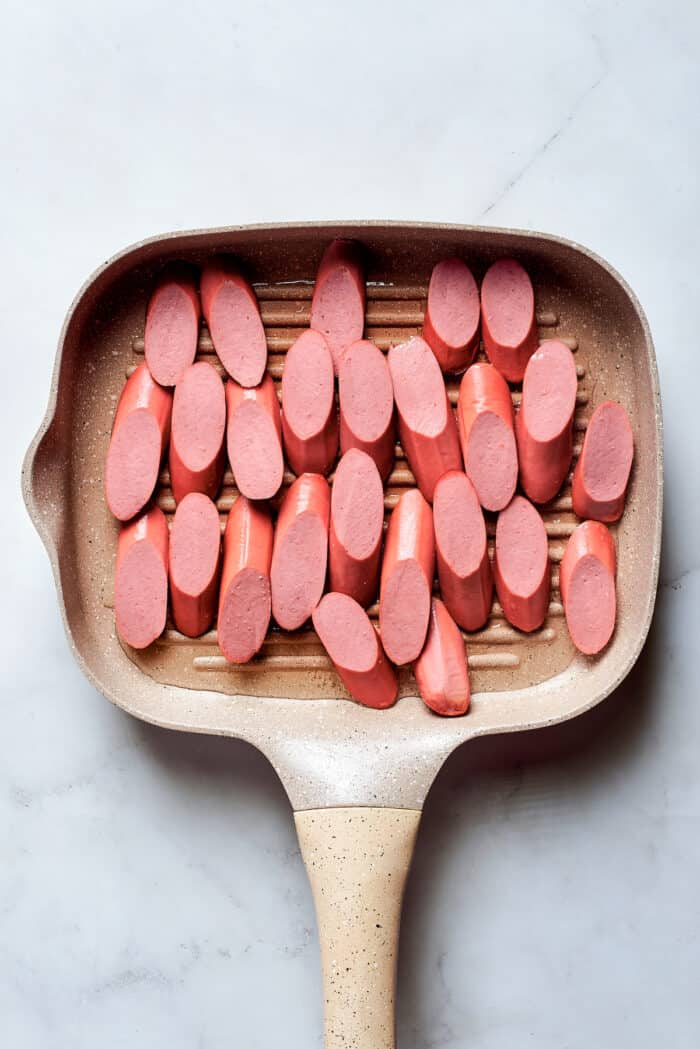 This Salchipapa recipe features crispy homemade french fries scattered with slices of beef hot dogs for a quick meal. A cast iron grill pan holds thick slices of hot dogs.