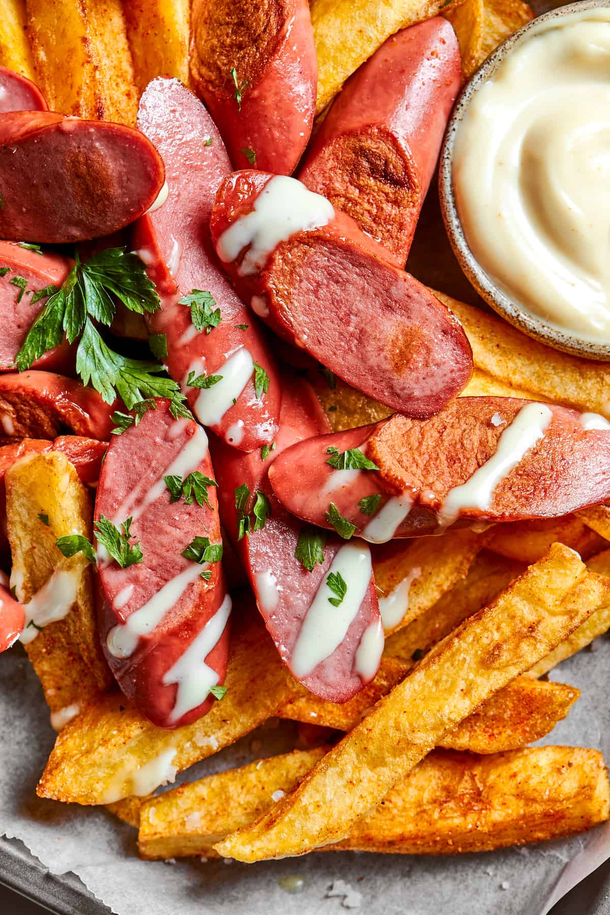 This Salchipapa recipe features crispy homemade french fries scattered with slices of beef hot dogs for a quick meal. A close up view of french fries and hot dog slices drizzled with mayonnaise and parsley, with a small bowl of mayo seen in the upper right.