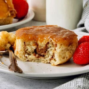 A caramel roll on a white plate with a piece cut out, a fork to its left, and strawberries to the right.