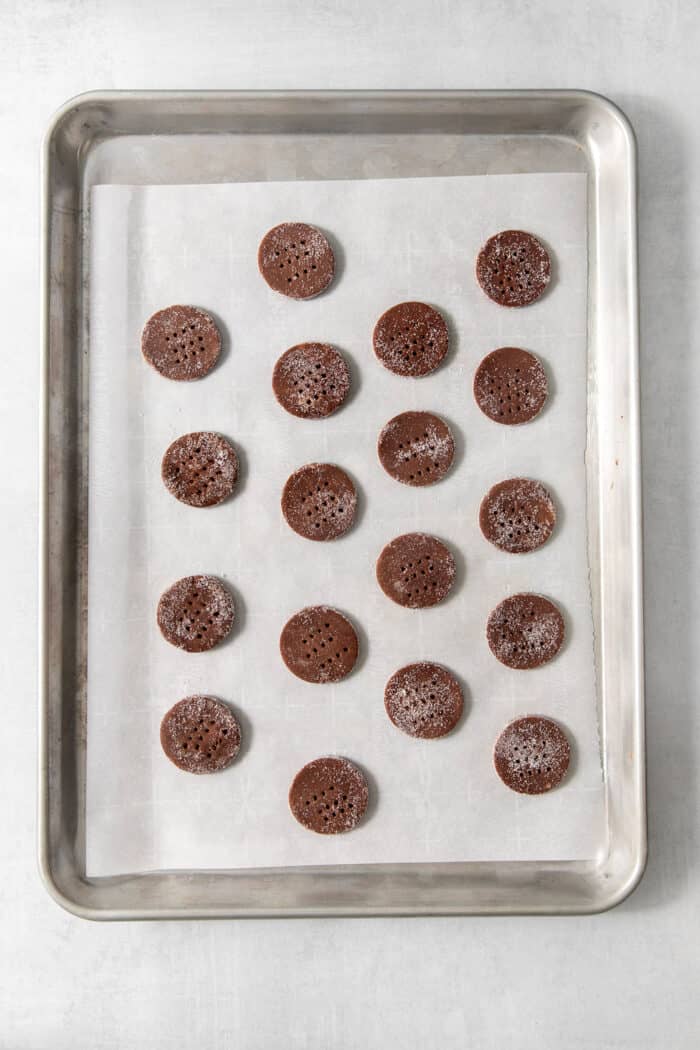 These Chocolate Cream Wafer Cookies feature buttery cocoa cookies that are soft, tender, and filled with a rich buttercream. Chocolate wafer cookie rounds are shown on a parchment-lined metal baking sheet.