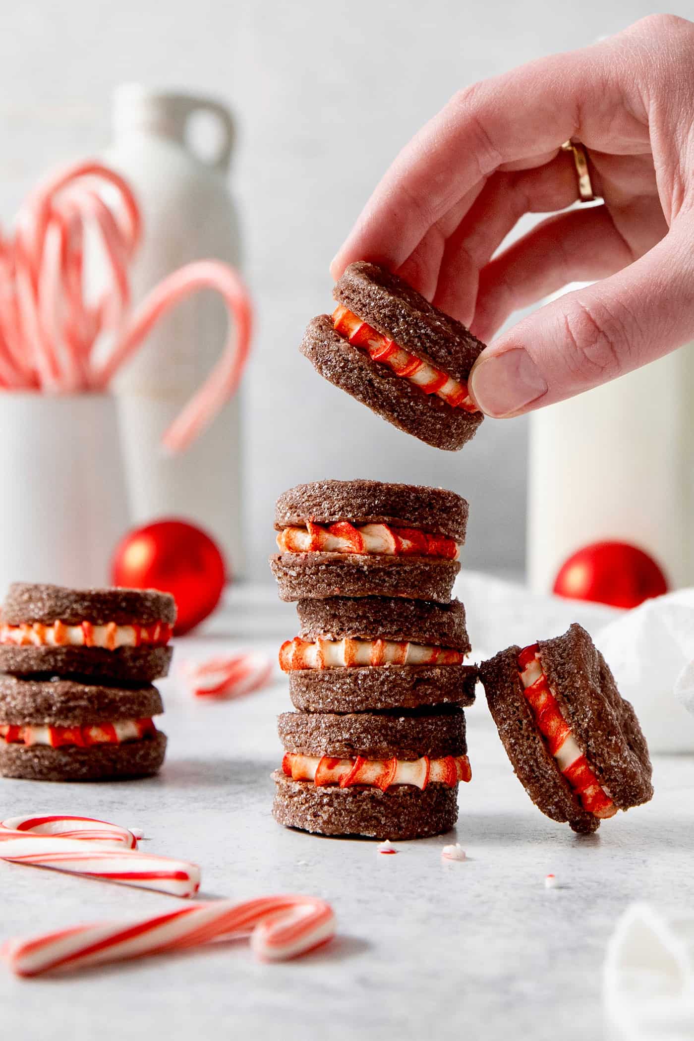 These Chocolate Cream Wafer Cookies feature buttery cocoa cookies that are soft, tender, and filled with a rich buttercream. A hand picks up a chocolate cream wafer cookie from a stack of cookies, with candy canes seen in the background.