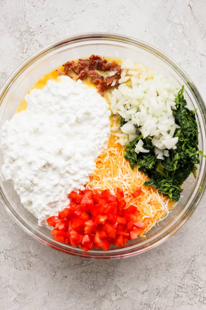 This protein-packed cottage cheese egg bake recipe combines creamy, fluffy eggs and cottage cheese with colorful veggies and crispy bacon. Ingredients are added to eggs in a glass bowl - pepper, flour, cheeses, bacon.