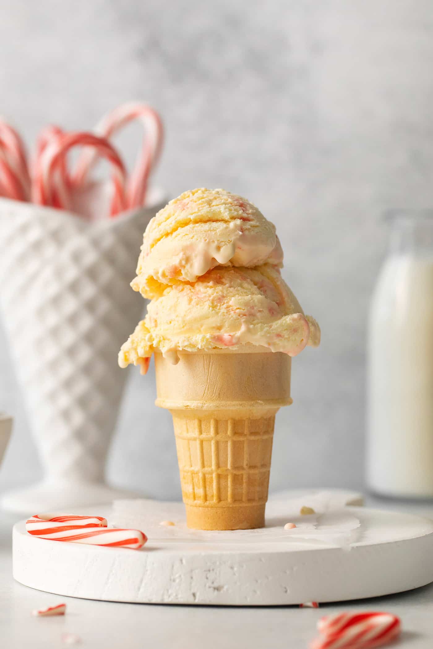 Creamy, dreamy Peppermint Ice Cream is so easy to make at home! Peppermint-infused ice cream is scattered with bits of candy canes. Two scoops of peppermint ice cream in an ice cream on a white marble round, with a ceramic cone of candy canes in the background.