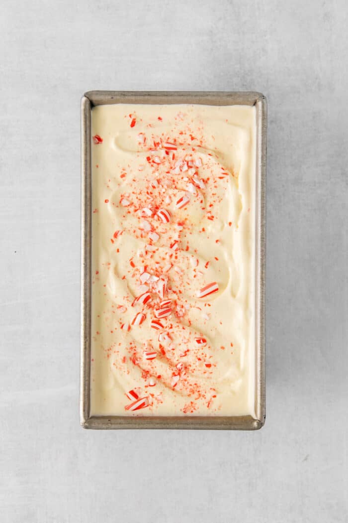 Creamy, dreamy Peppermint Ice Cream is so easy to make at home! Peppermint-infused ice cream is scattered with bits of candy canes. A metal loaf pan holds cream-colored ice cream topped with crushed candy cane pieces.
