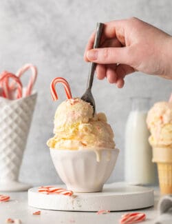 A hand holding a spoon digs into a bowl containing two scoops of peppermint ice cream topped by a candy cane.
