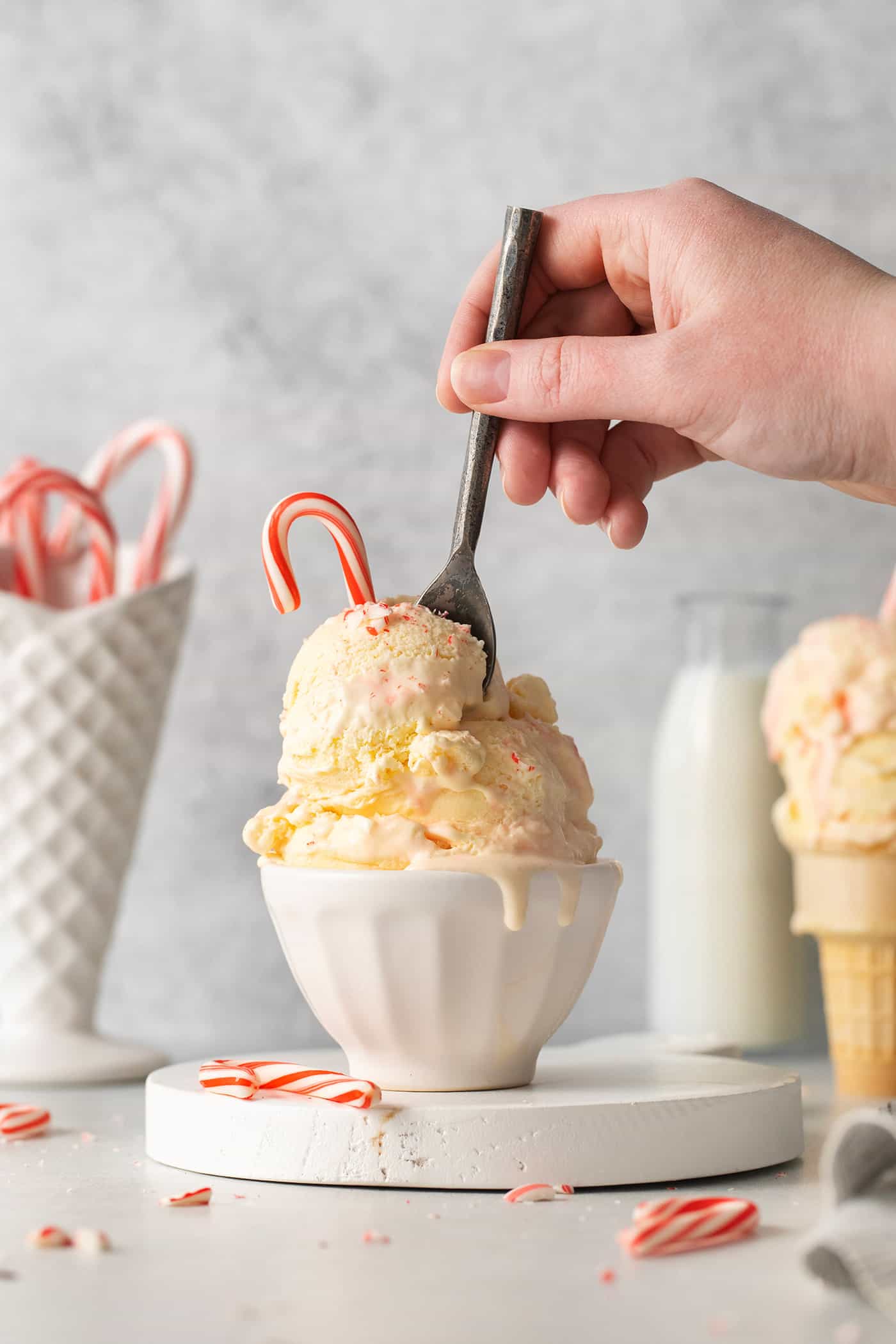 Creamy, dreamy Peppermint Ice Cream is so easy to make at home! Peppermint-infused ice cream is scattered with bits of candy canes. A hand holding a spoon digs into a bowl containing two scoops of peppermint ice cream topped by a candy cane.