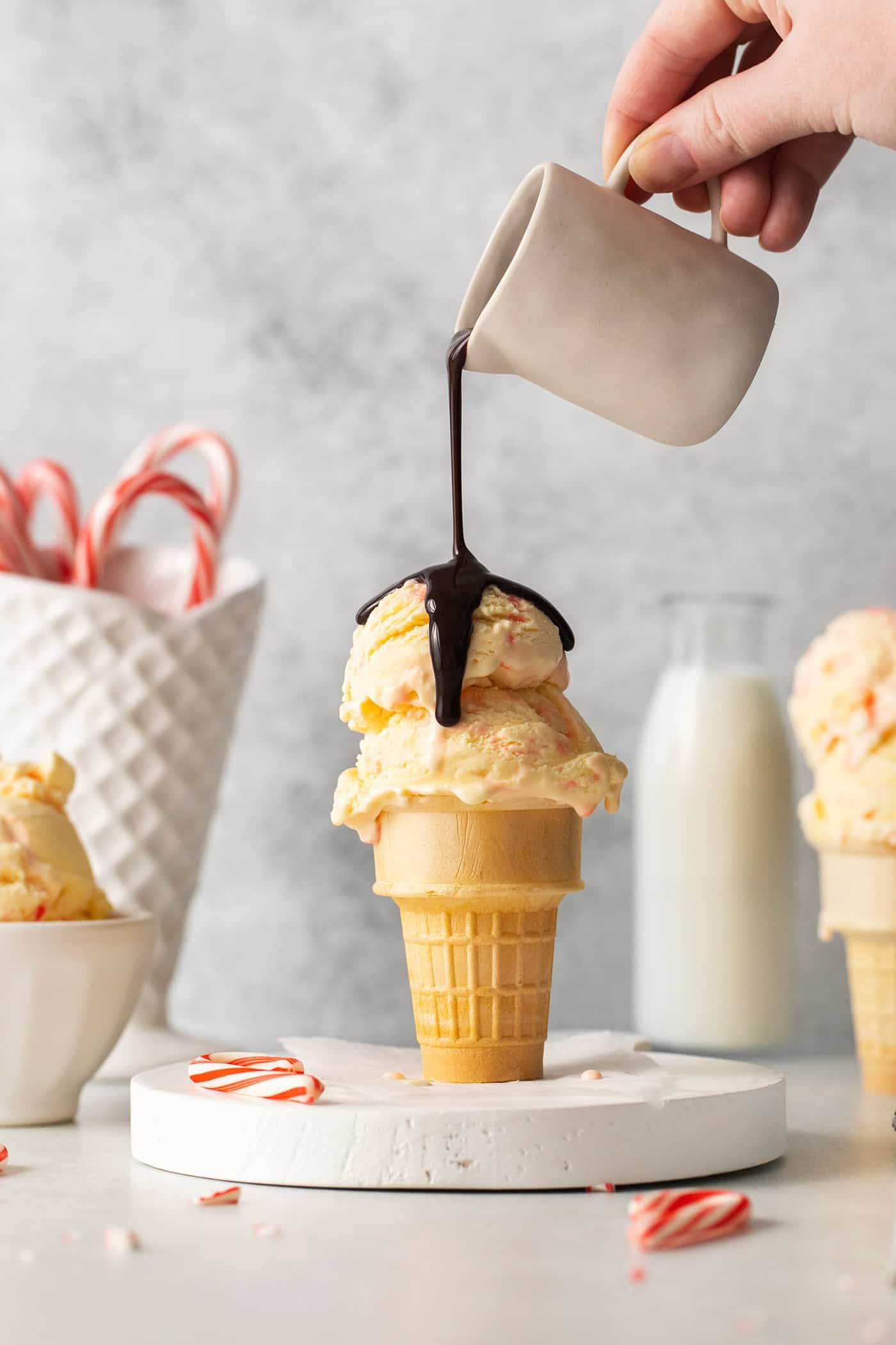 Creamy, dreamy Peppermint Ice Cream is so easy to make at home! Peppermint-infused ice cream is scattered with bits of candy canes. A hand holding a white pitcher pours chocolate sauce over an ice cream cone full of two scoops of peppermint ice cream, with candy canes in the background.