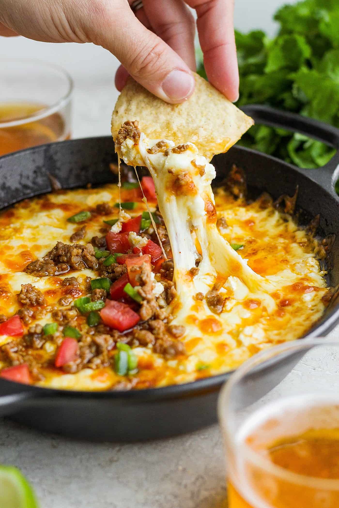 Creamy and just a little spicy, Queso Fundido is an easy, go-to dip! Melted cheese and chorizo are topped with fresh tomatoes and jalapeno. A hand holding a corn chip dips into a bowl of tomato-topped queso fundido with melted cheese and chorizo.