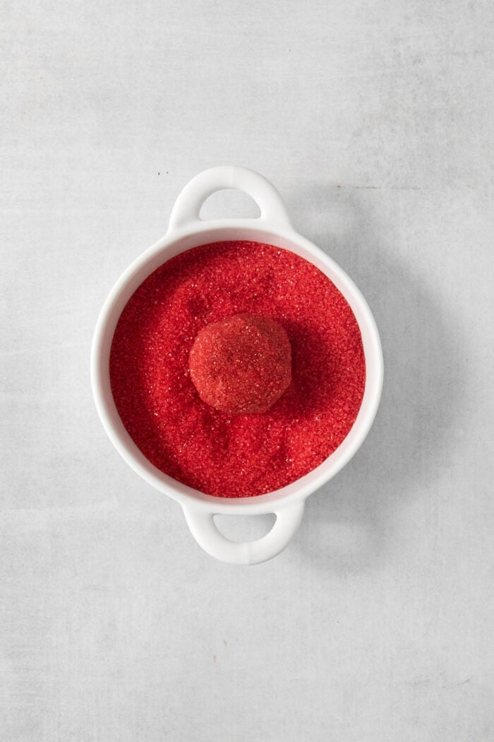 These Red Velvet Cookies are soft and chewy, with a crisp sugar coating and a hint of cocoa and vanilla for flavor. A red velvet cookie is placed in a bowl of red sanding sugar.
