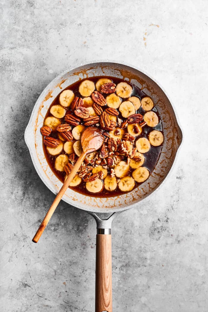 Bananas Foster French Toast features rich and tender slices of egg-soaked sweet bread that are topped with rum-spiked caramelized bananas. A wooden spoon stirs a skillet of cooking banana slices for bananas foster.
