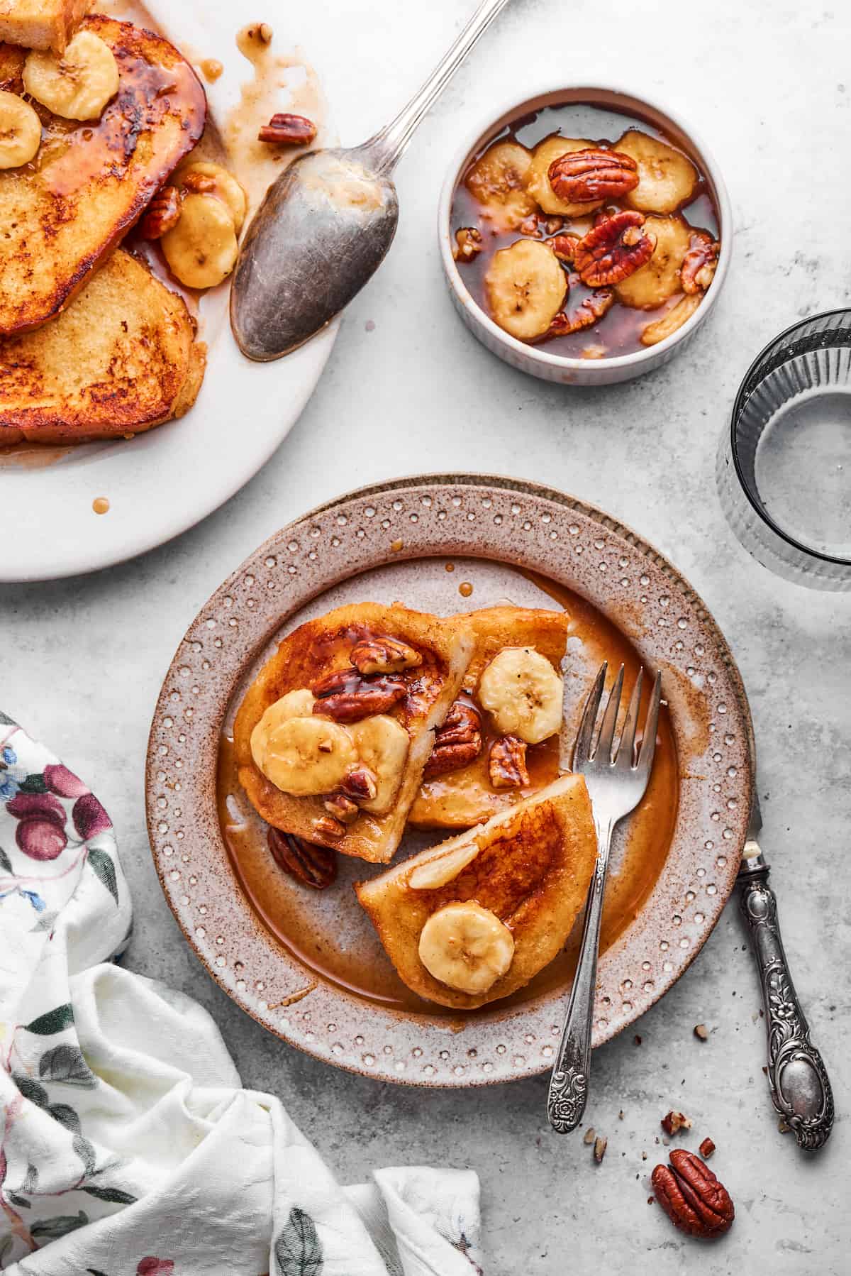 Bananas Foster French Toast features rich and tender slices of egg-soaked sweet bread that are topped with rum-spiked caramelized bananas. A plate of bananas foster French toast with a fork and a small bowl of bananas in sauce nearby, with more french toast to the left.