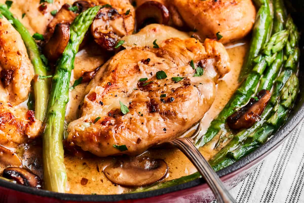 Creamy Chicken Madeira pairs tender chicken breasts with bright asparagus in a rich sauce. Add savory sautéed mushrooms and sweet wine. A serving utensil lifts out a piece of chicken from a creamy pan of chicken madeira.