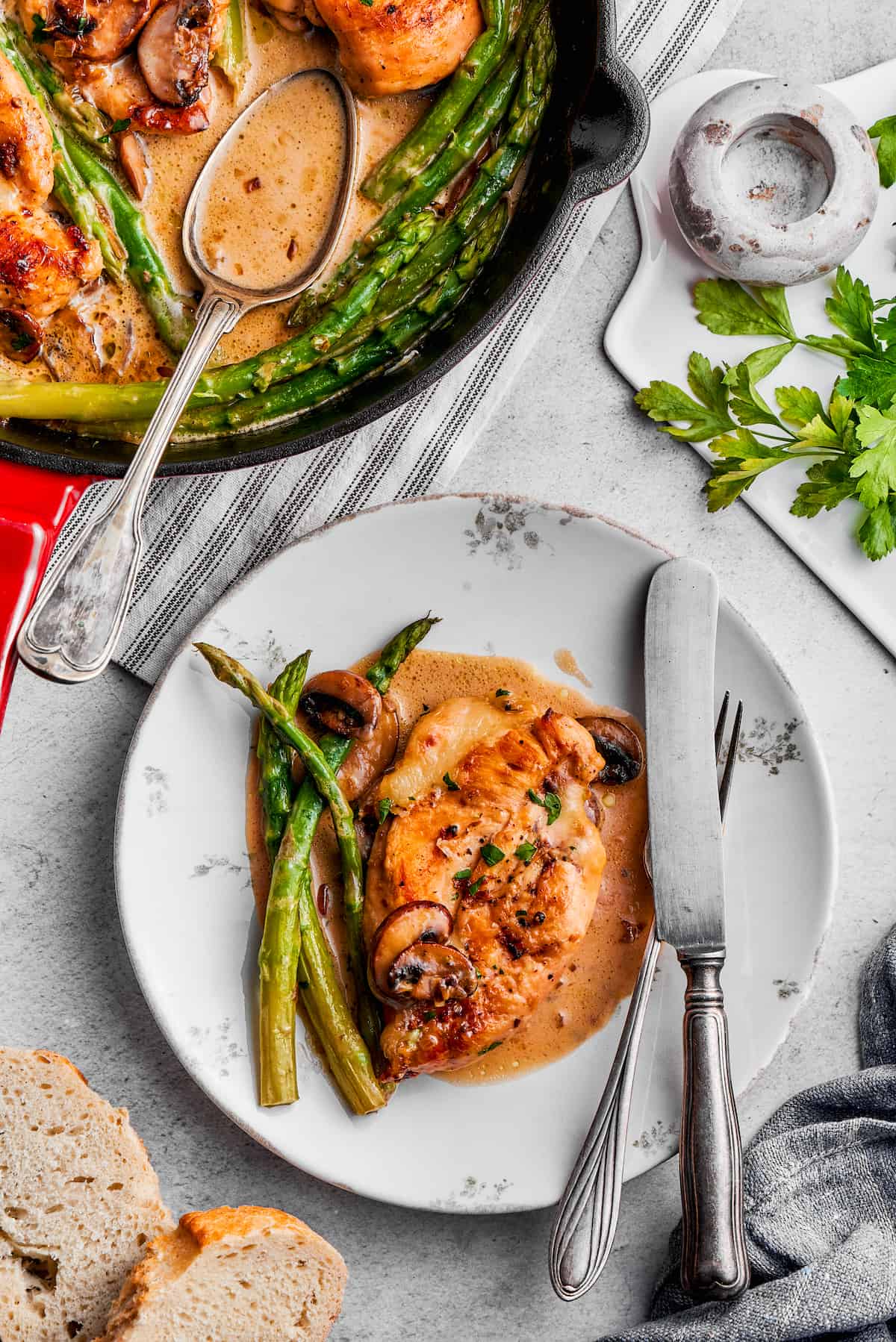 Creamy Chicken Madeira pairs tender chicken breasts with bright asparagus in a rich sauce. Add savory sautéed mushrooms and sweet wine. A plate of chicken madeira with knife and fork resting on its edge, next to a pan of chicken madeira.