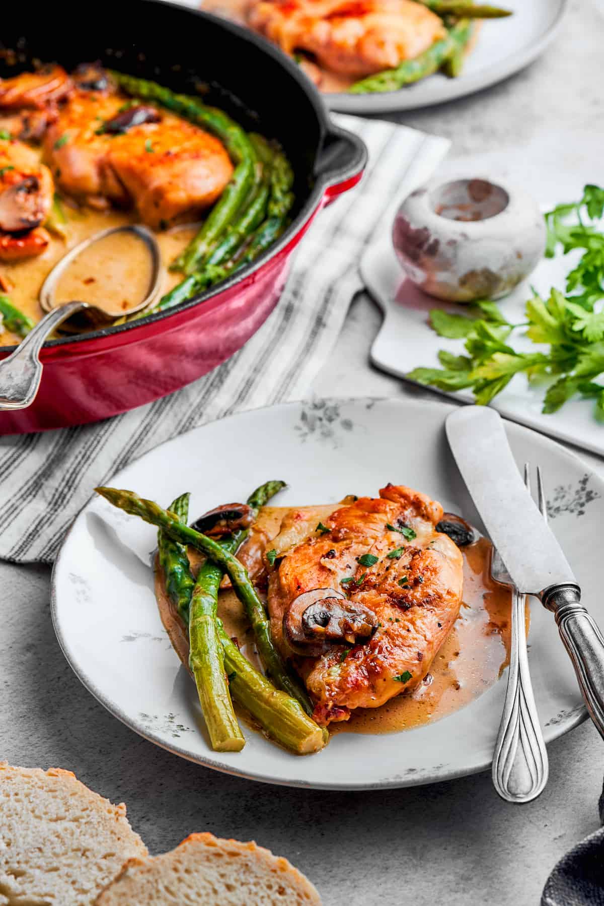 Creamy Chicken Madeira pairs tender chicken breasts with bright asparagus in a rich sauce. Add savory sautéed mushrooms and sweet wine. A table view of a plate of chicken madeira with utensils and the pan of chicken.