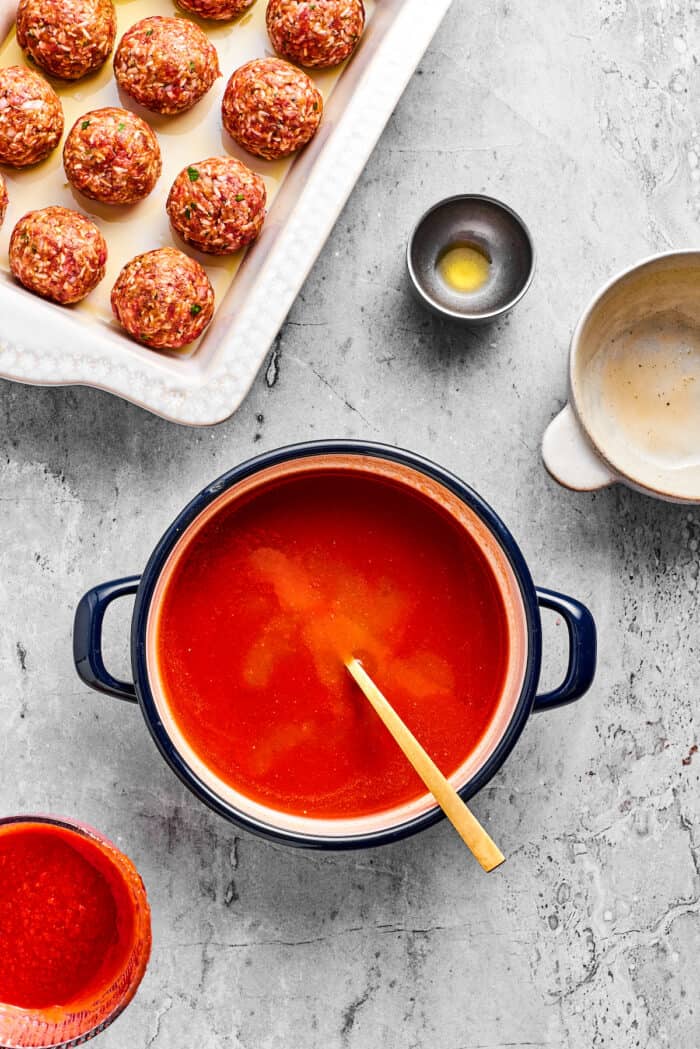 Tender beef and rice Porcupine Meatballs are smothered in tomato sauce to make a savory, filling dish that's perfect with mashed potatoes. A wooden spoon stirs a pot of tomato sauce with uncooked porcupine meatballs to its left.