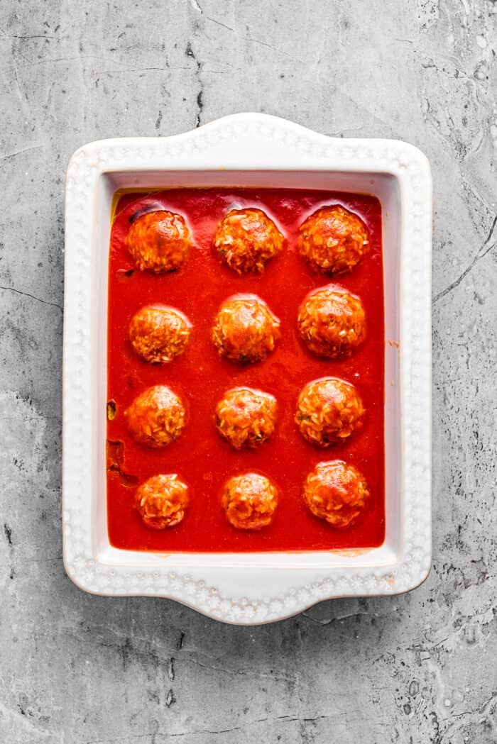 Tender beef and rice Porcupine Meatballs are smothered in tomato sauce to make a savory, filling dish that's perfect with mashed potatoes. Meatballs are placed in red sauce in a white baking dish.