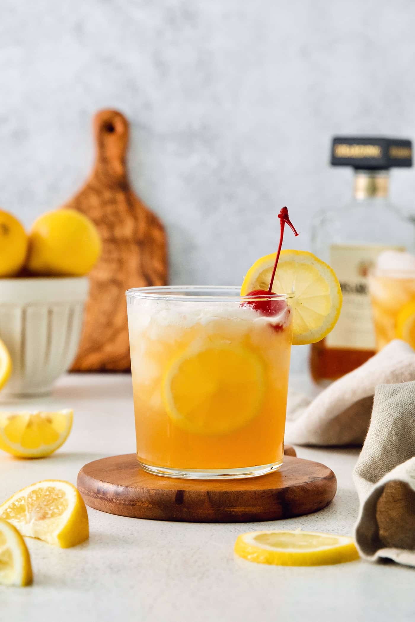 This Amaretto Sour is a classic cocktail featuring sweet almond-flavored amaretto liqueur and tart fresh lemon juice for a flavorful drink. A lemon-garnished glass of amaretto sour is shown on a wooden circle with lemon slices around it and lemons in the background.