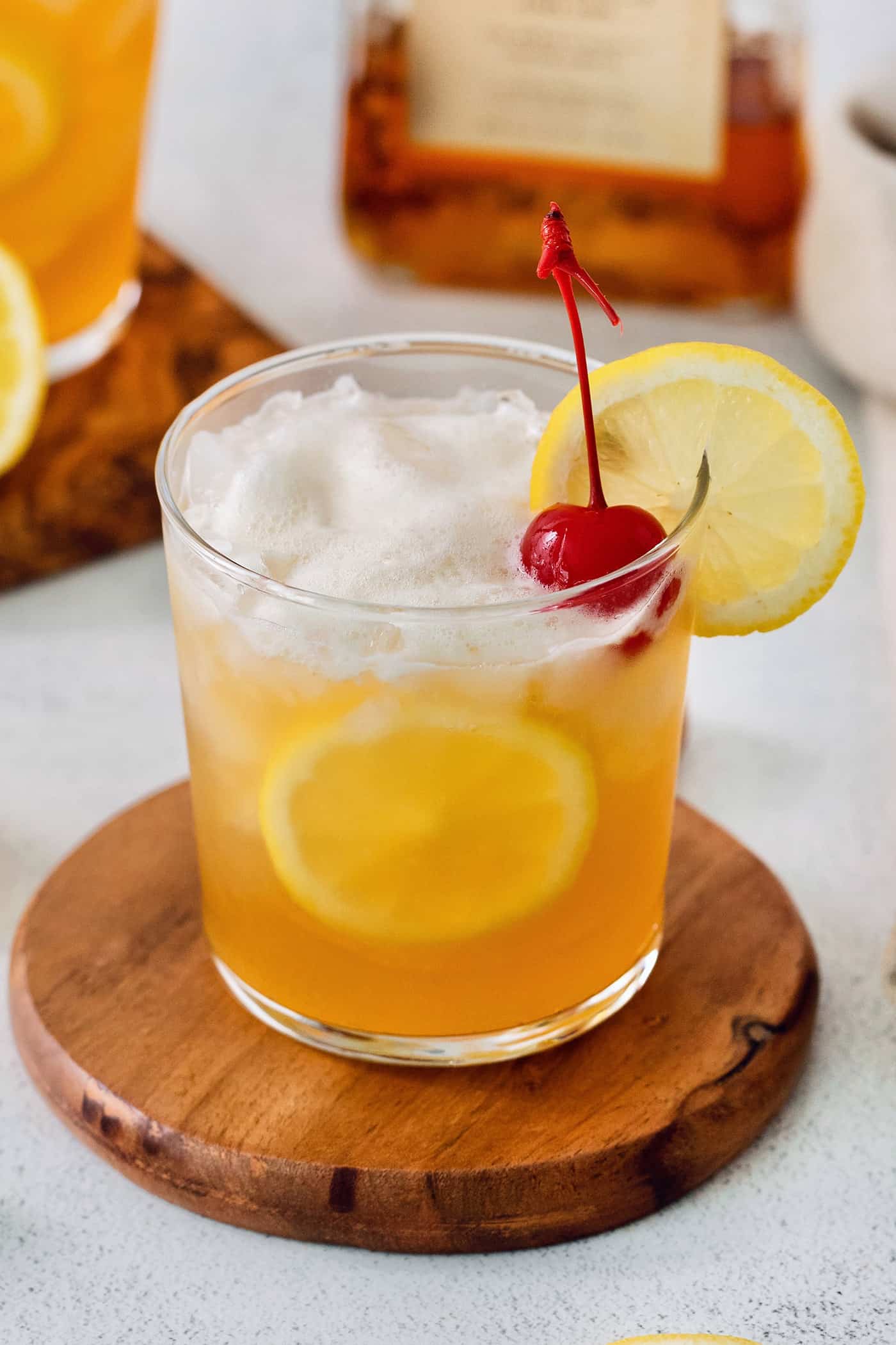This Amaretto Sour is a classic cocktail featuring sweet almond-flavored amaretto liqueur and tart fresh lemon juice for a flavorful drink. A glass of amaretto sour is garnished with a cherry and a slice of lemon on a wooden circle.