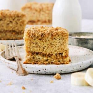 A piece of banana coffee cake on a white plate with a fork, with more pieces of cake shown in the background.