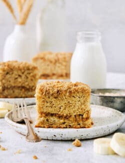 A piece of banana coffee cake on a white plate with a fork, with more pieces of cake shown in the background.