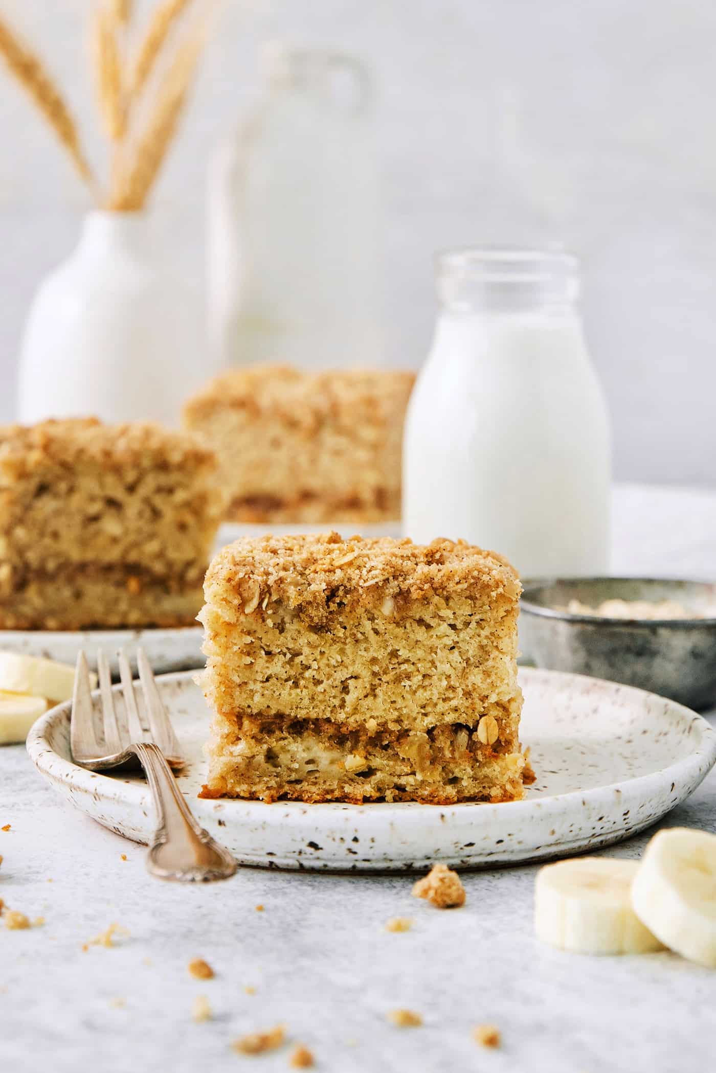 This Banana Coffee Cake features a tender banana bread cake layered with an oat streusel and topped with an optional vanilla bean glaze. A piece of banana coffee cake on a white plate with a fork, with more pieces of cake shown in the background.