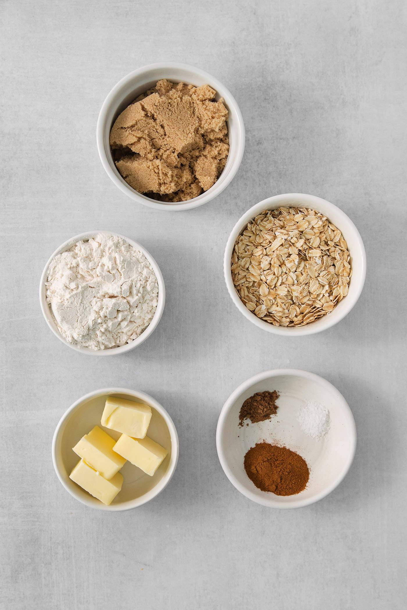 This Banana Coffee Cake features a tender banana bread cake layered with an oat streusel and topped with an optional vanilla bean glaze. The ingredients for banana coffee cake streusel are shown portioned out in bowls: butter, flour, oats, brown sugar, salt, cinnamon, and nutmeg.