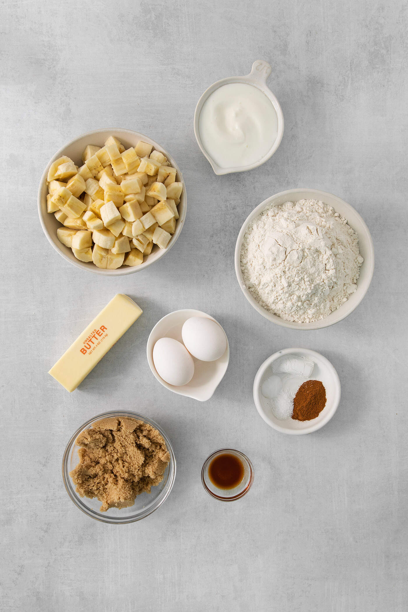 This Banana Coffee Cake features a tender banana bread cake layered with an oat streusel and topped with an optional vanilla bean glaze. The ingredients for banana coffee cake are shown portioned out in bowls: butter, flour, eggs, vanilla, brown sugar, sour cream, cinnamon, baking powder, baking soda, and salt, and bananas,