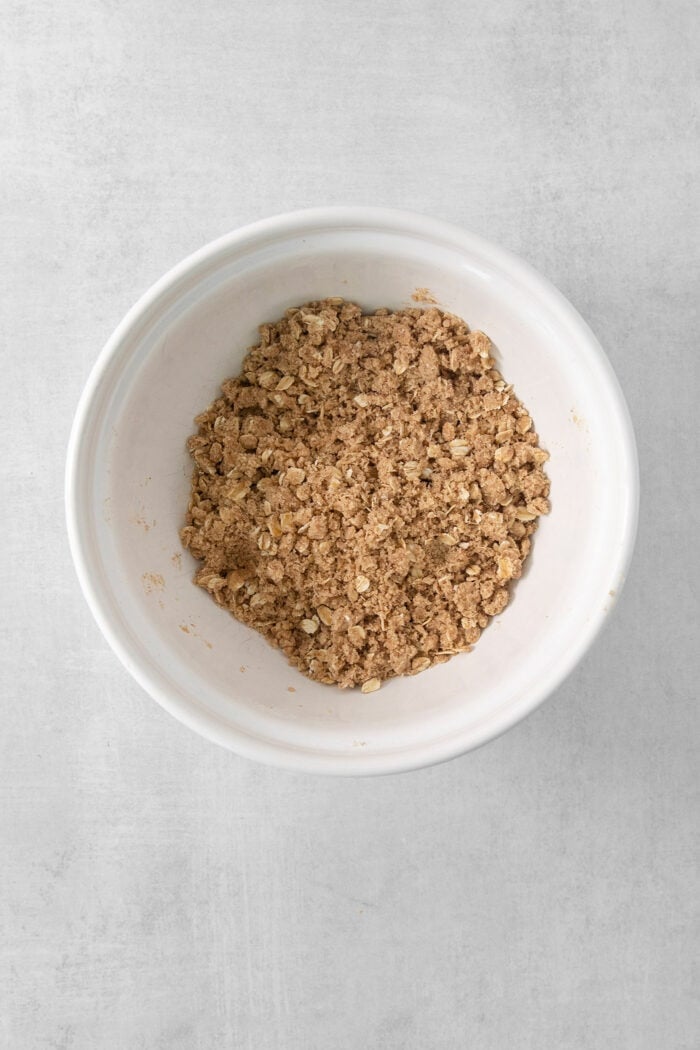 This Banana Coffee Cake features a tender banana bread cake layered with an oat streusel and topped with an optional vanilla bean glaze. A white bowl of streusel for banana coffee cake.