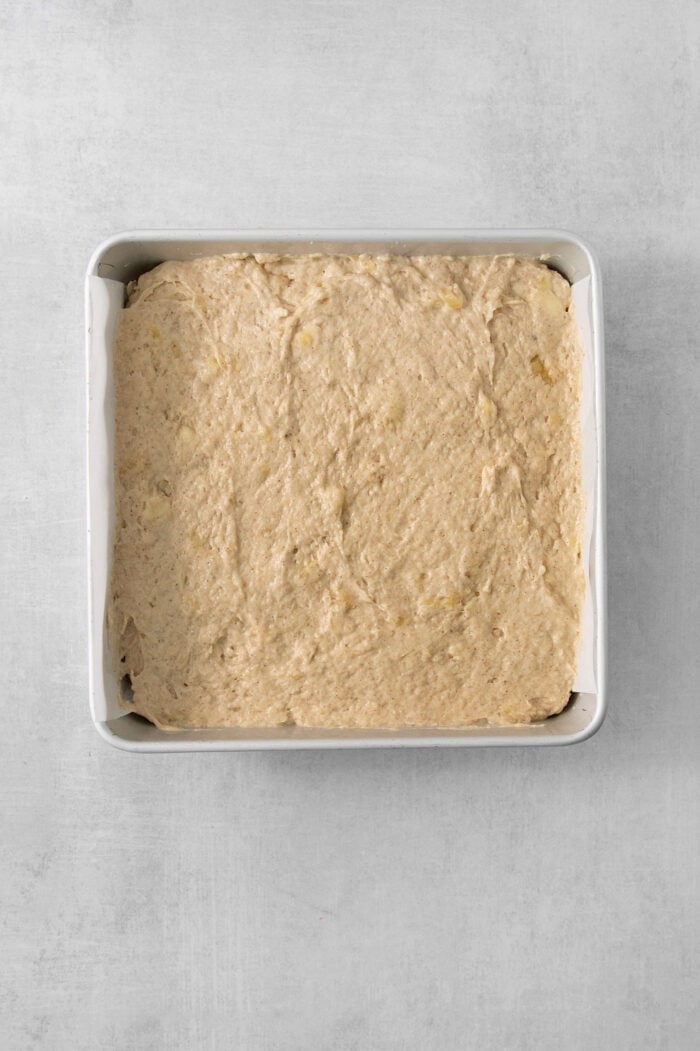 This Banana Coffee Cake features a tender banana bread cake layered with an oat streusel and topped with an optional vanilla bean glaze. The batter for banana coffee cake is spread out in a metal baking pan.