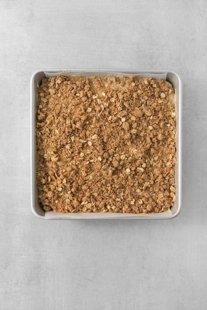 This Banana Coffee Cake features a tender banana bread cake layered with an oat streusel and topped with an optional vanilla bean glaze. A metal baking pan of streusel-topped banana coffee cake on a white background.