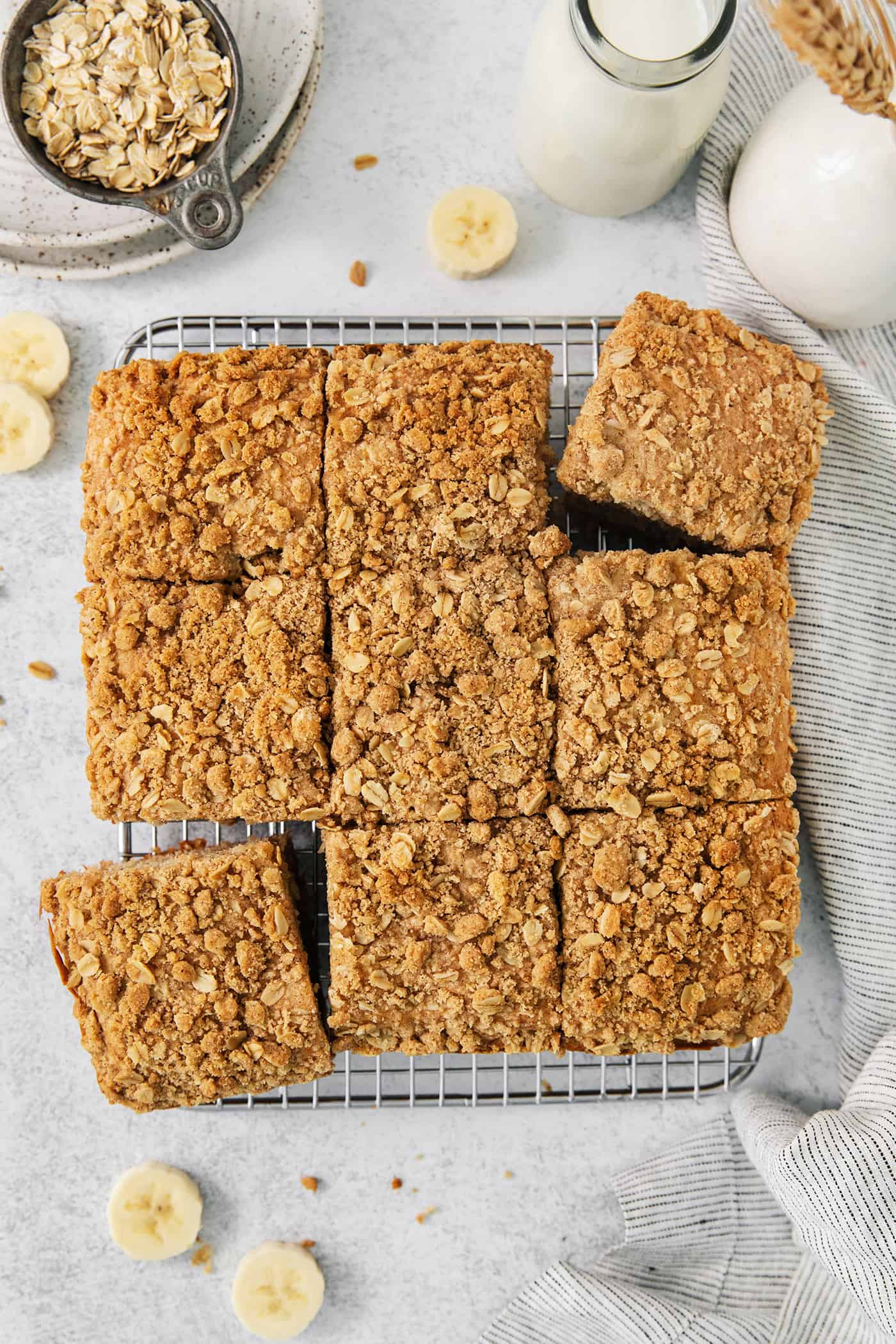 This Banana Coffee Cake features a tender banana bread cake layered with an oat streusel and topped with an optional vanilla bean glaze. Banana coffee cake is cut into 9 squares with banana slices scatted around them.