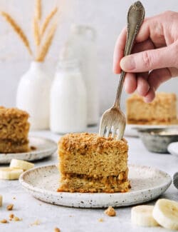 A hand holding a fork cuts into a thick slice of banana coffee cake on a white plate.