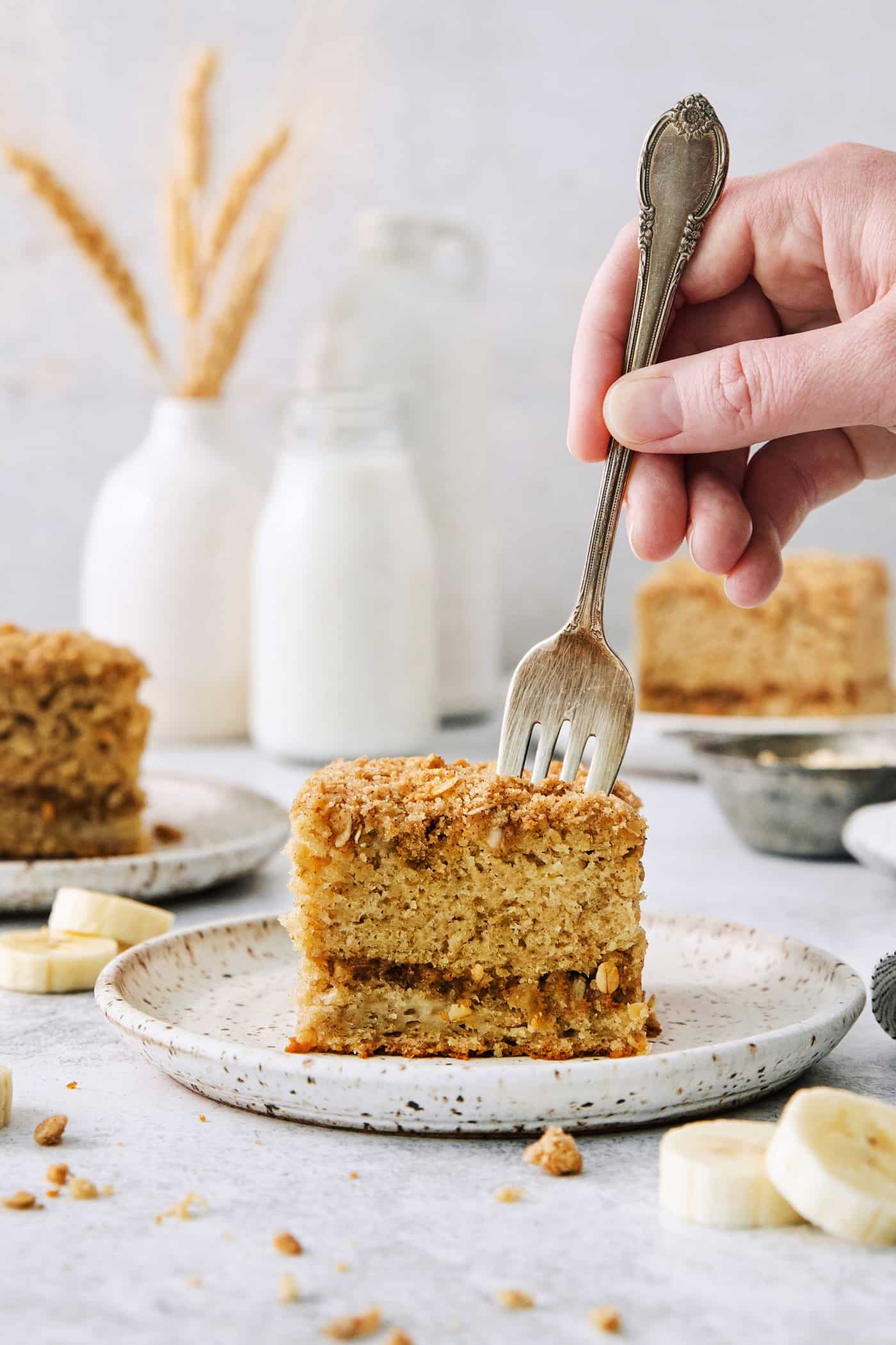 This Banana Coffee Cake features a tender banana bread cake layered with an oat streusel and topped with an optional vanilla bean glaze. A hand holding a fork cuts into a thick slice of banana coffee cake on a white plate.