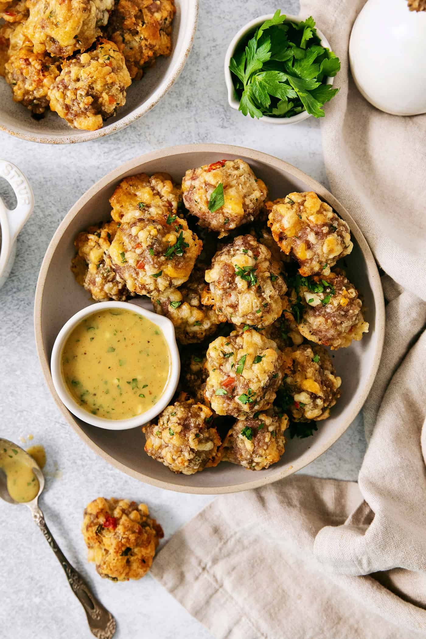 A big white bowl with serving utensils holds sausage cheese balls with a bowl of yellow dipping sauce nestled against the balls.
