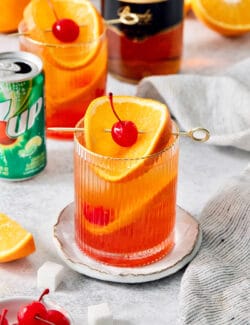 Glasses of brandy alexander garnished with orange and cherries, with a can of 7UP in the background.