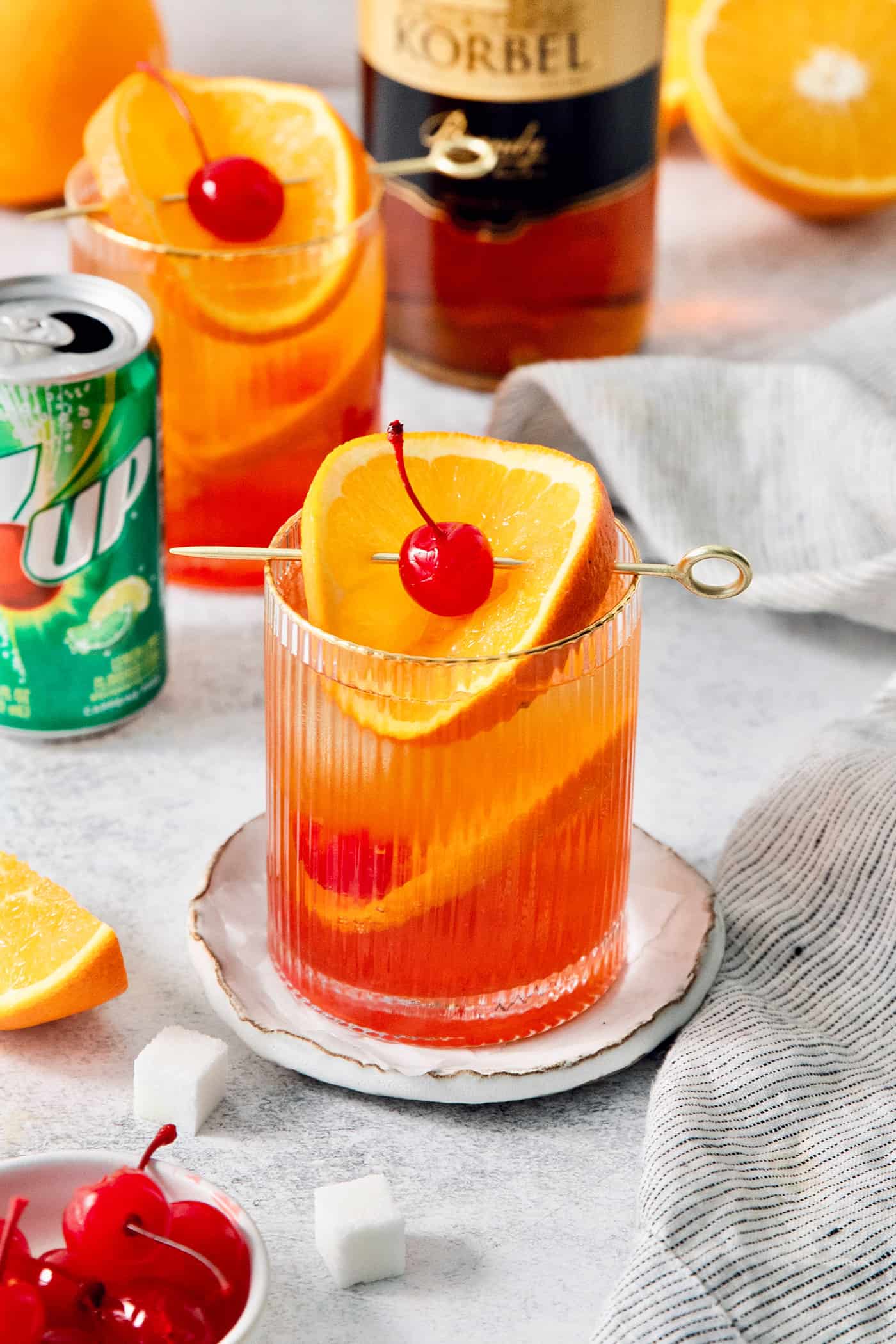 Glasses of brandy alexander garnished with orange and cherries, with a can of 7UP in the background.